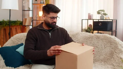 Indian man on sofa opens box and shakes head after discovering wrong item in 스톡 동영상 347033359