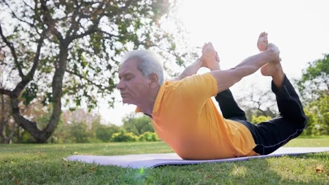 Indian senior citizen man doing Dhanurasana Yoga outdoors in morning sunlight. Stock Footage 249941626
