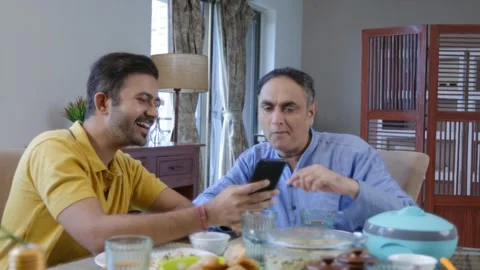 Indian son at dining table showing funny video on mobile phone to his old dad. Stock Footage 217427038