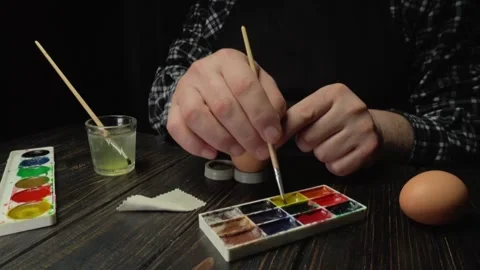 Individual decorates an egg with paint using a brush, surrounded by watercolor Stock Footage 330407537
