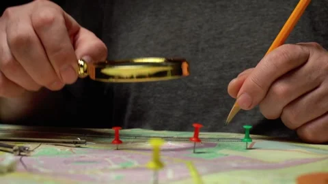 Individual examines a map with a magnifying glass while marking locations with Stock Footage 332830404