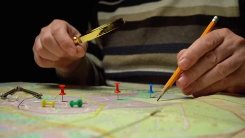 Individual examines a map with a magnifying glass while using a pencil to mark Stock Footage 333308039