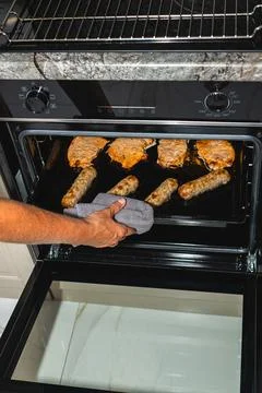 Individual with a gray oven mitt pulls out sausages and chicken from a black Stock Photos