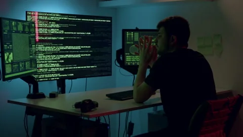 Individual seated at a desk with multiple computer monitors displaying code and Video stock 314270577