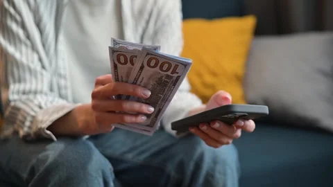 An individual is sitting on a couch and counting cash while using a smartphone. Video stock 338446102