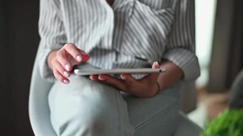 Individual uses a tablet while sitting on a chair indoors. The hands are Stockbeeldmateriaal 336778560