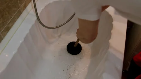 Individual using a black plunger to clear a clogged bathtub drain, demonstrating Stock Footage 325481945