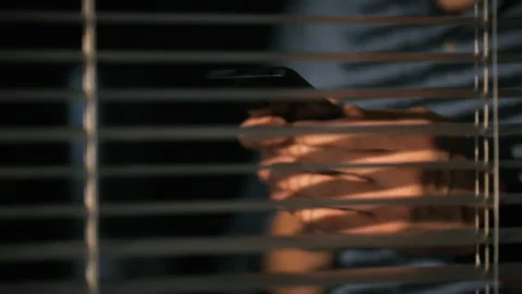 Individual using a smartphone while hidden behind closed blinds Stock Footage 304277296