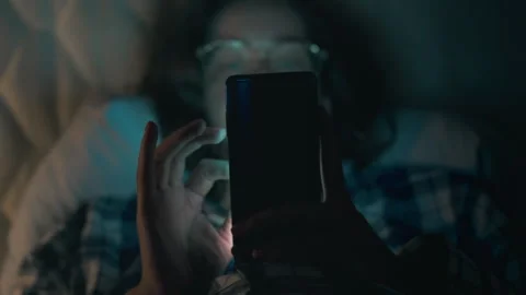 Individual using a smartphone while lying in bed during evening hours at hom Stock Footage 323709721
