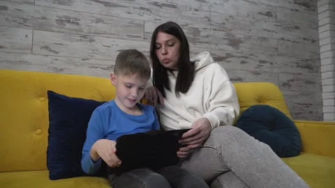 Indoors, sitting on a yellow sofa mom watches her son play on a tablet helps him Stock Footage 257515288