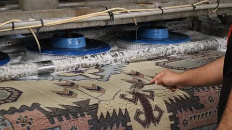Industrial brush machine cleaning patterned wool rug while worker applies Foto stock