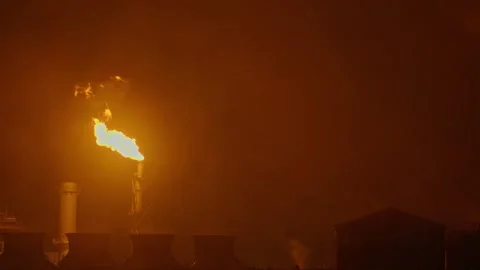 An industrial flare stack burns off excess gas with a large, bright orange flame Stock Footage 322073707