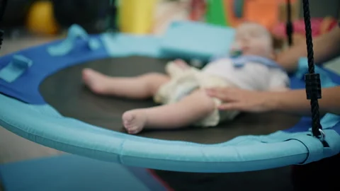 Infant lying on a trampoline during an early intervention session, therapist's 스톡 동영상 342870198