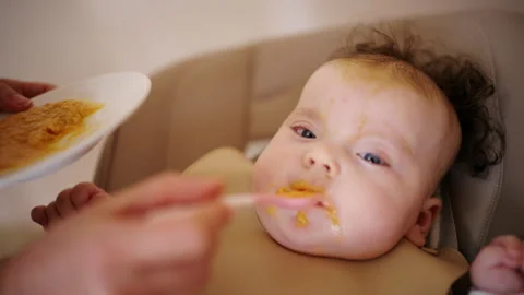 Infant seated in a high chair while an adult feeds the baby with a spoon Stock Footage 329398492
