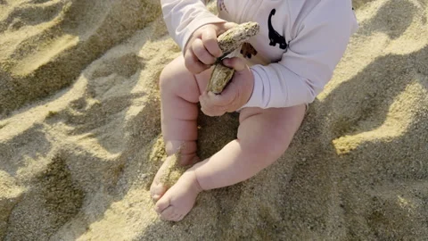 Infant sitting on sandy beach, holding two stones, wearing light pink outfit Stock Footage 313948286
