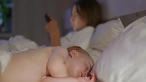 Infant sleeping peacefully on bed while mother uses smartphone in dimly lit Stock-Footage 341897492