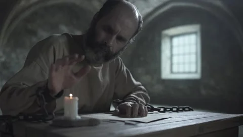 Inmate in shackles pausing his writing, warming hand over a burning candle Stock Footage 341009396