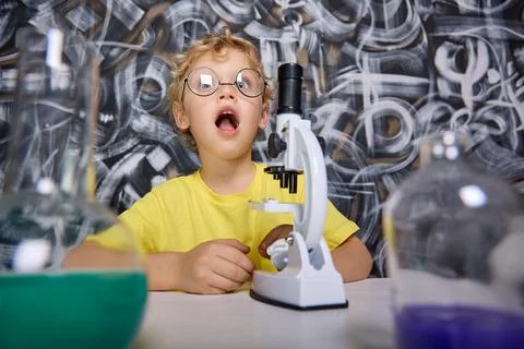 Inquisitive boy sits at table with microscope opened his mouth wide in surprise Stock Photos