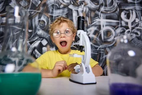 Inquisitive boy sits at table with microscope opened his mouth wide in surprise Stock Photos