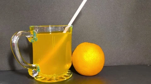 Inserting straw into a clear glass cup of orange juice next to a whole oran.. Video stock 318126164