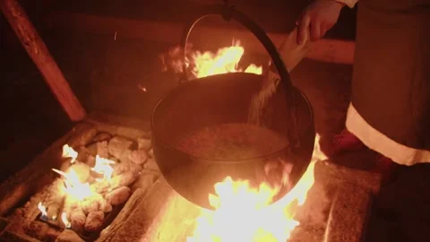 Inside a rustic kitchen, a large black kettle simmers over a fireplace while a Stock Footage 336428520