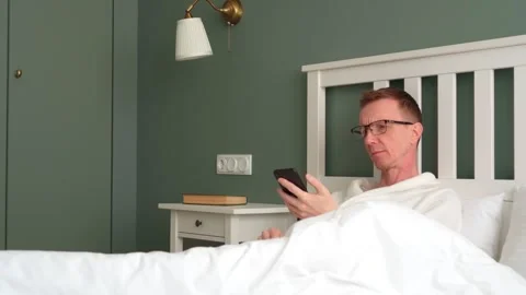 Insomnia. Man with reading glasses is lying in bed and looking at information on Stock Footage 260336904