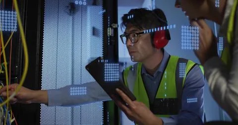 Inspecting Asian tech using tablet and leaning into server rack in data center, Foto stock