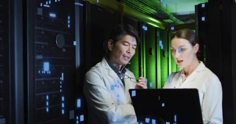 Inspecting servers, techs in white lab coats viewing laptop in data center aisle Stock Photos