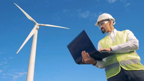 Inspector in warnvest is observing a functioning windmill with his laptop Video stock 97998412