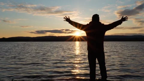 An inspired man raises his hands up, standing on the shore of the lake at sunset Stock Footage 275083648