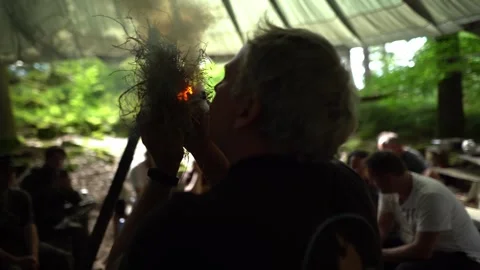 Instructor blowing on glowing ember inside natural tinder bundle to ignite Stock Footage 330970298