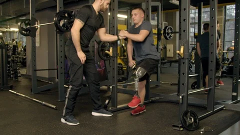 Instructor giving a hand to young man exercising with prosthetic leg in gym Stock Footage 125531122
