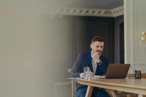 Intellectual bearded man makes notes in notebook concentrated at laptop screen Stock Photos