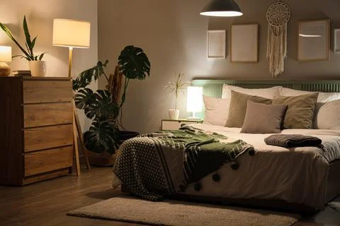 Interior of bedroom with blankets on bed, plants and glowing lamps at night Stock Photos