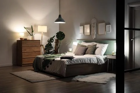Interior of bedroom with blankets on bed, plants and glowing lamps at night Stock Photos