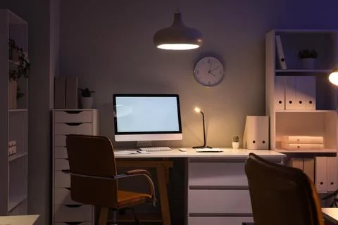 Interior of dark office with workplace, shelf units and glowing lamps at nigh Stock Photos