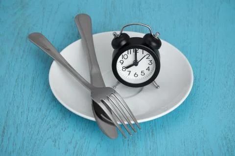 Intermittent fasting concept with clock on white plate, fork and knife on blu Stock Photos