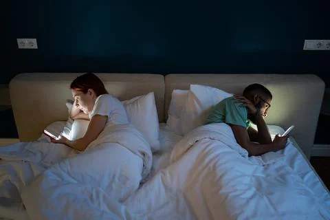 Interracial couple lying back to back in bed, using smartphones, feeling Stock-Fotos