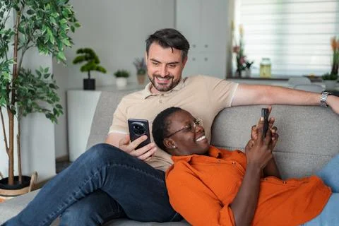 Interracial couple relaxing on sofa using smartphones at home Stock Photos