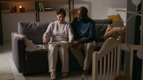 Interracial Couple Sorting Through Baby Clothes on Couch in Nursery Stock Footage 319965501
