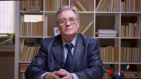 Interview of senior businessman talking into camera on book shelves background. Video stock 108413095
