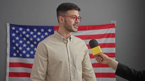 Interviewee man in glasses speaking into microphone against a usa flag back.. 스톡 동영상 271098460
