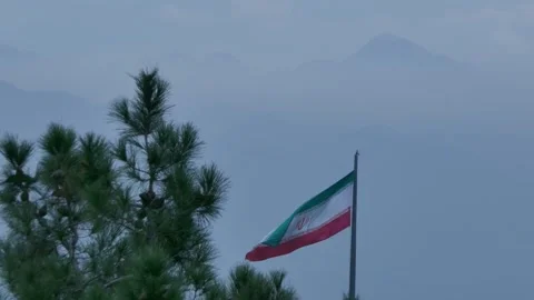 Iranian flag waving beside pine trees with misty mountain backdrop Stock Footage 317836621