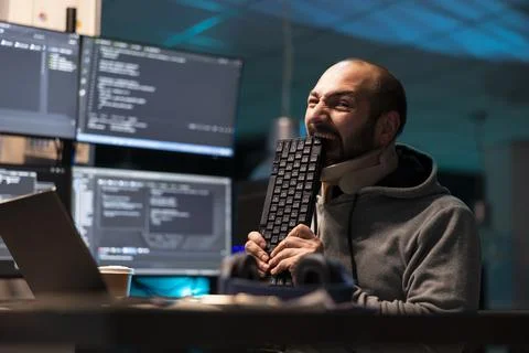 Irritated male coder biting keyboard at office desk 库存照片