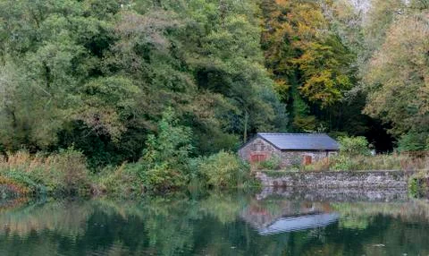 An isolated, low stone building, reflected in a calm lake, with trees behind it Foto stock