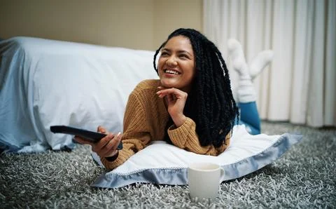 Its time for some comedy. a young woman lying on the floor with a remote control Stock Photos