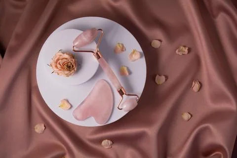 Jade roller, gua sha and dry rose and petals on a white podium. Stock Photos