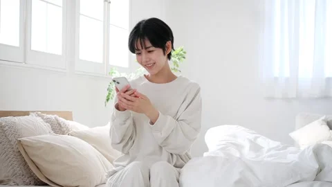 Japanese Woman Using Smartphone While Relaxing in Bright Bedroom at Home 스톡 동영상 343839619