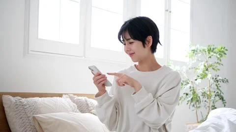Japanese Woman Using Smartphone While Relaxing in Bright Bedroom at Home Stock-Footage 343840137
