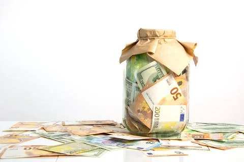 A jar on a white surface holds both dollar and euro bills, with a brown lid.  Stock Photos